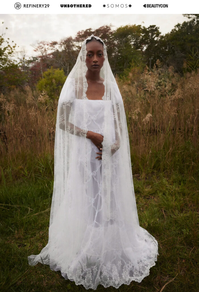 A woman in a white lace veil and dress stands in a grassy field, looking directly at the camera.