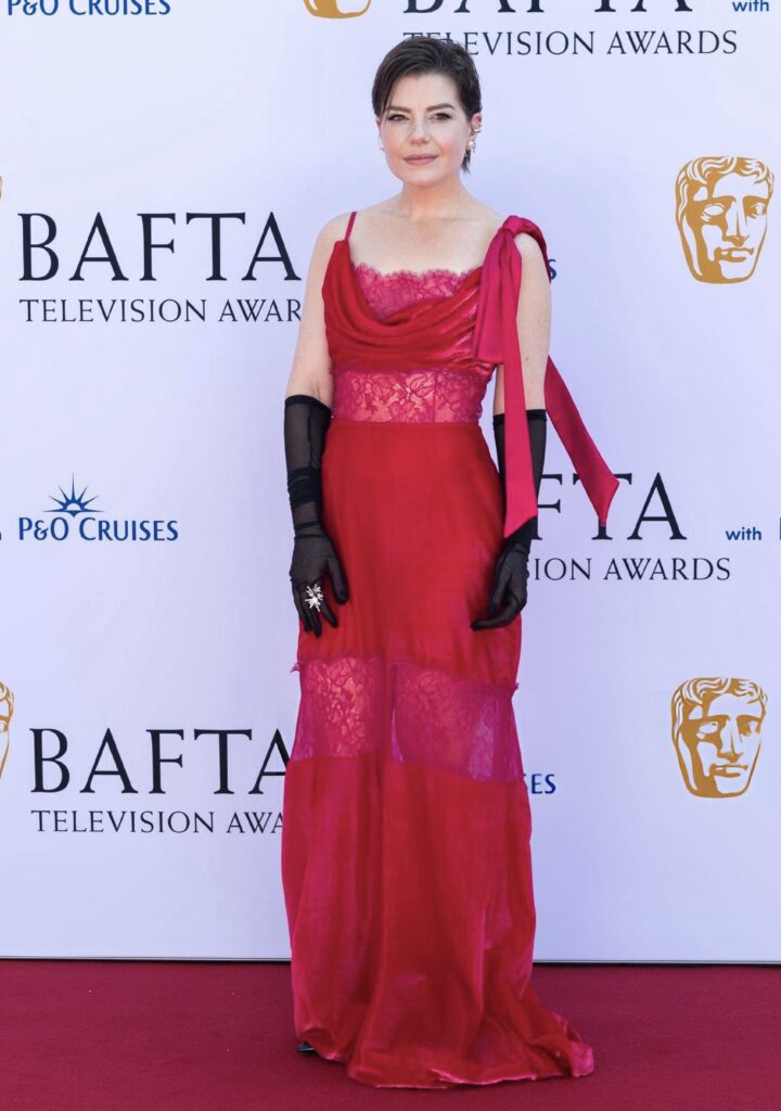 Woman in a red lace gown with black gloves stands on a red carpet at the BAFTA Television Awards.