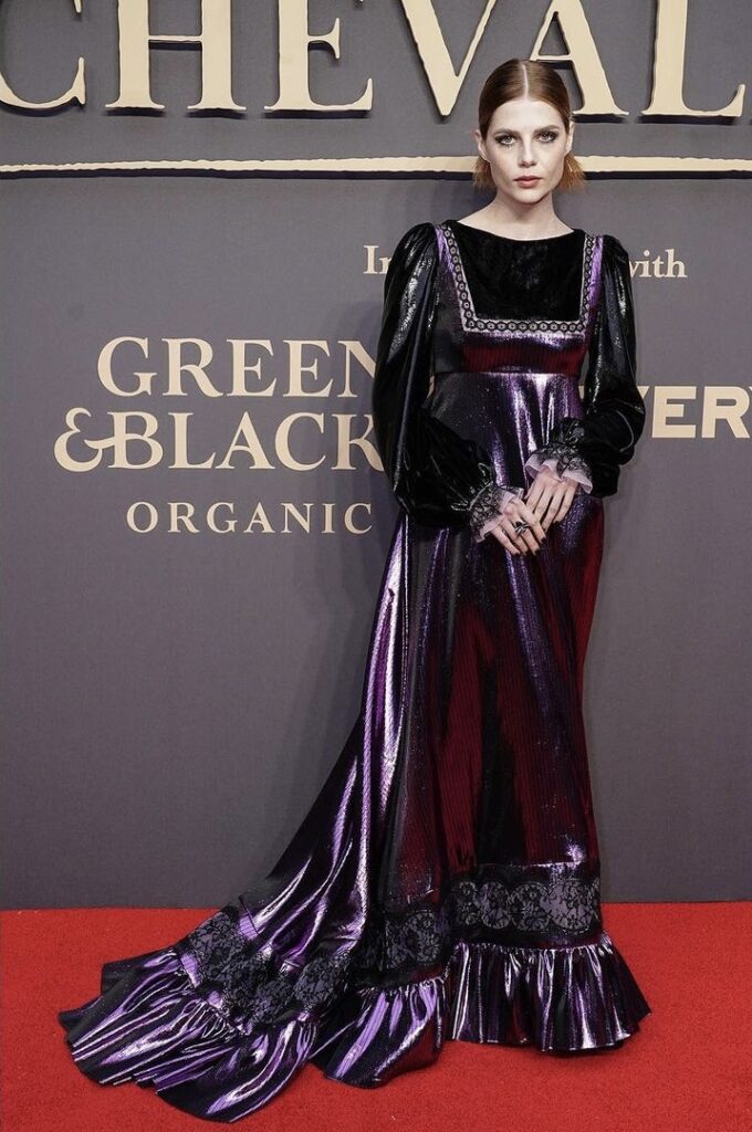 Woman in a shiny purple and black gown with lace details on a red carpet in front of an event backdrop.