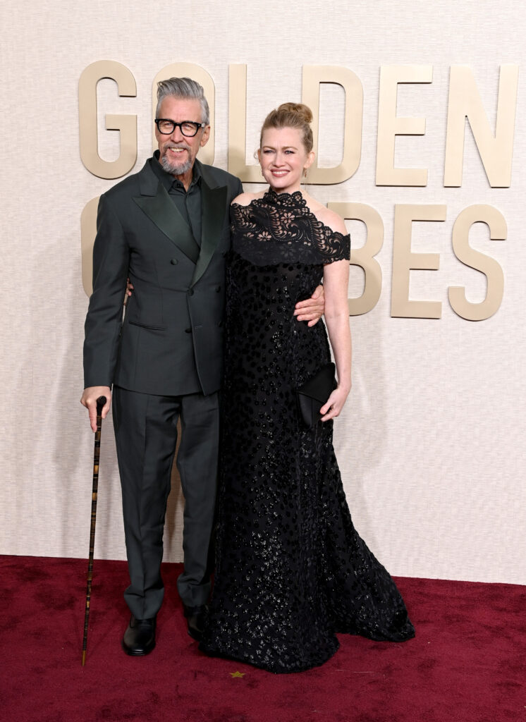 A man in a dark suit with a cane and a woman in a black gown pose on a red carpet at an event.