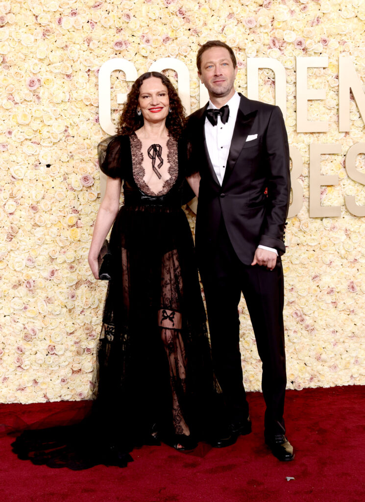 A woman in a black dress and a man in a tuxedo pose together on a red carpet with a floral backdrop.