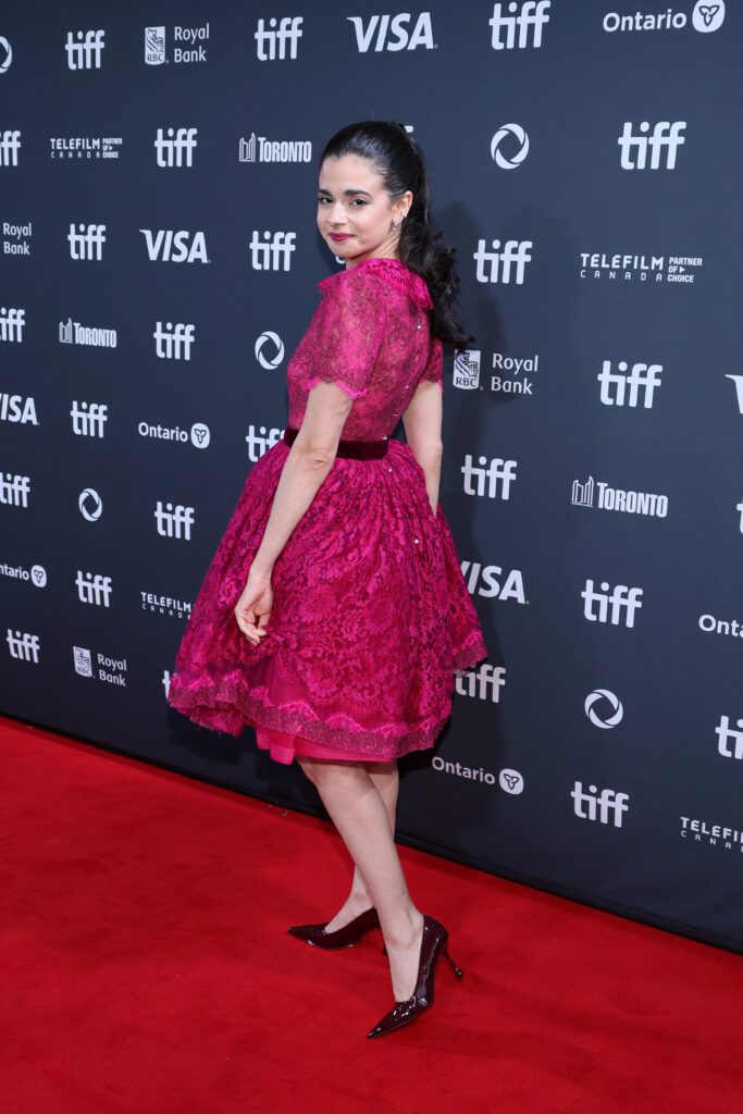 Woman in a pink lace dress poses on the red carpet at a film festival event, facing sideways and smiling.