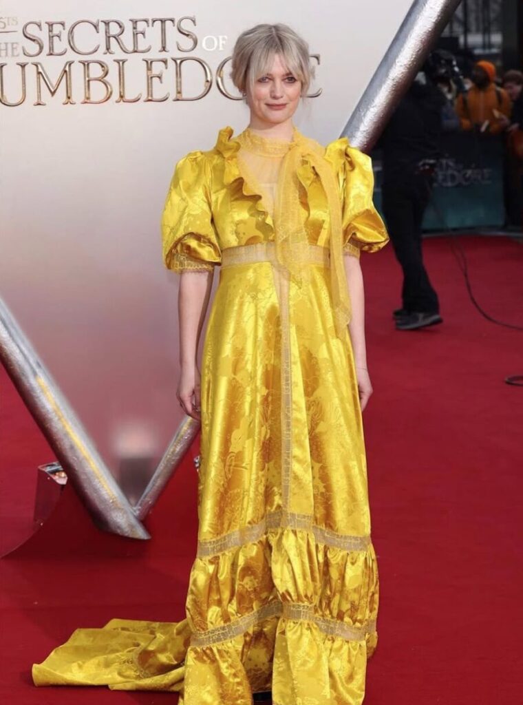 Person in a bright yellow, ruffled gown stands on a red carpet at a movie premiere event.