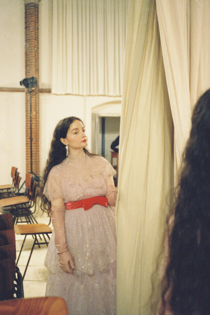A woman in a pink, glittery dress with a red belt stands by a curtain in a room with empty chairs.