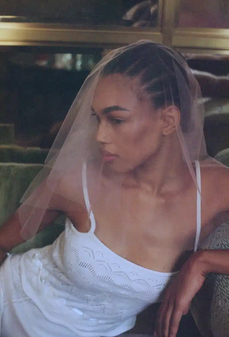 A woman wears the Embroidered Heart Veil and a white dress as she sits on a green couch, gazing thoughtfully to the side.