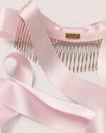 The Satin Bow Hair Comb, featuring a pink satin ribbon on a metal comb, rests on a pale background.