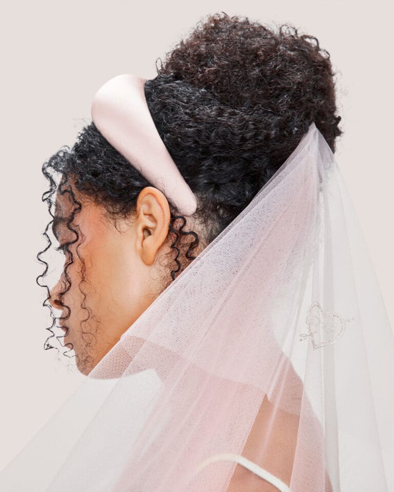 Woman with curly hair wearing the Embroidered Heart Veil in Pink tulle with the Pink Padded Satin Headband attachment, photographed in front of a light pink background.