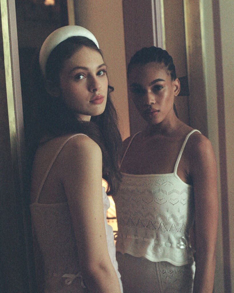 Two women in Cashmere Heart Knit outfits stand closely together looking toward the camera in soft, dim lighting.