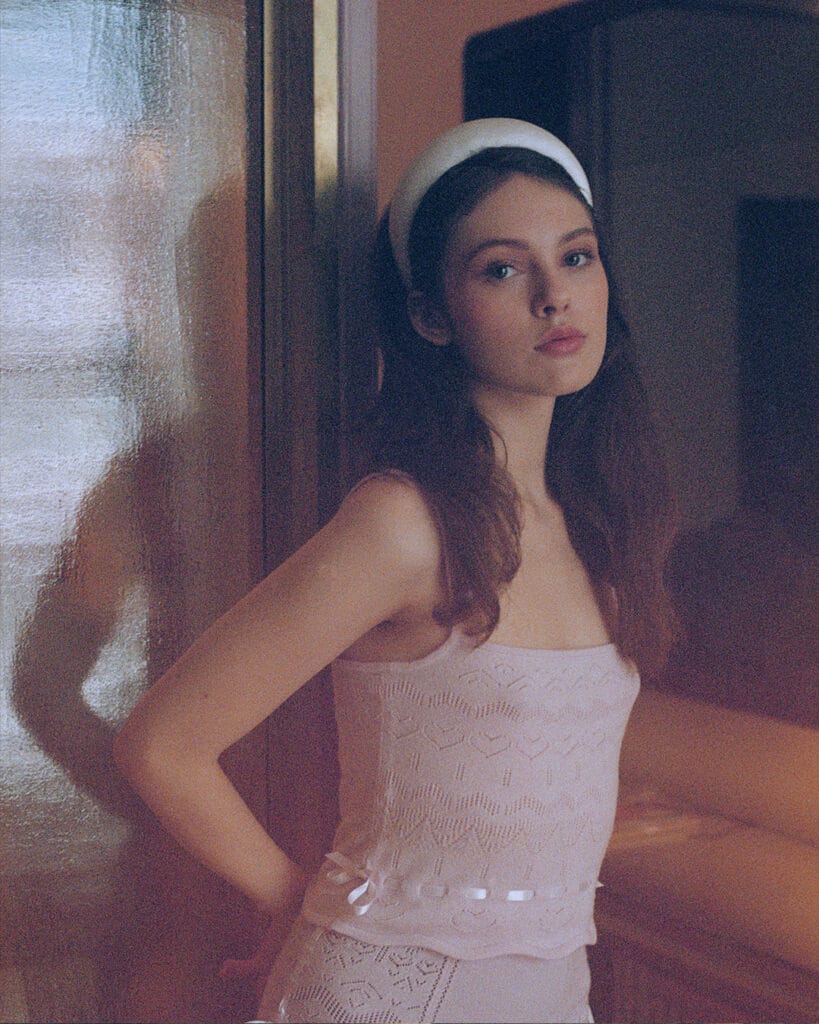 A woman with long hair wears the Cashmere Heart Knit Camisole and a t White Padded Satin Headband, standing in front of a window.