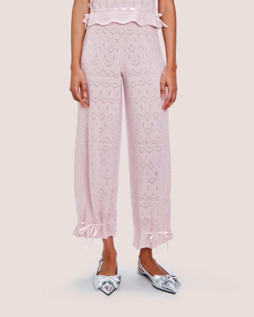 A woman wears Cashmere Heart Knit Pants in light pink with lace detailing and ribbon ties at the ankles, paired with silver pointed-toe shoes.