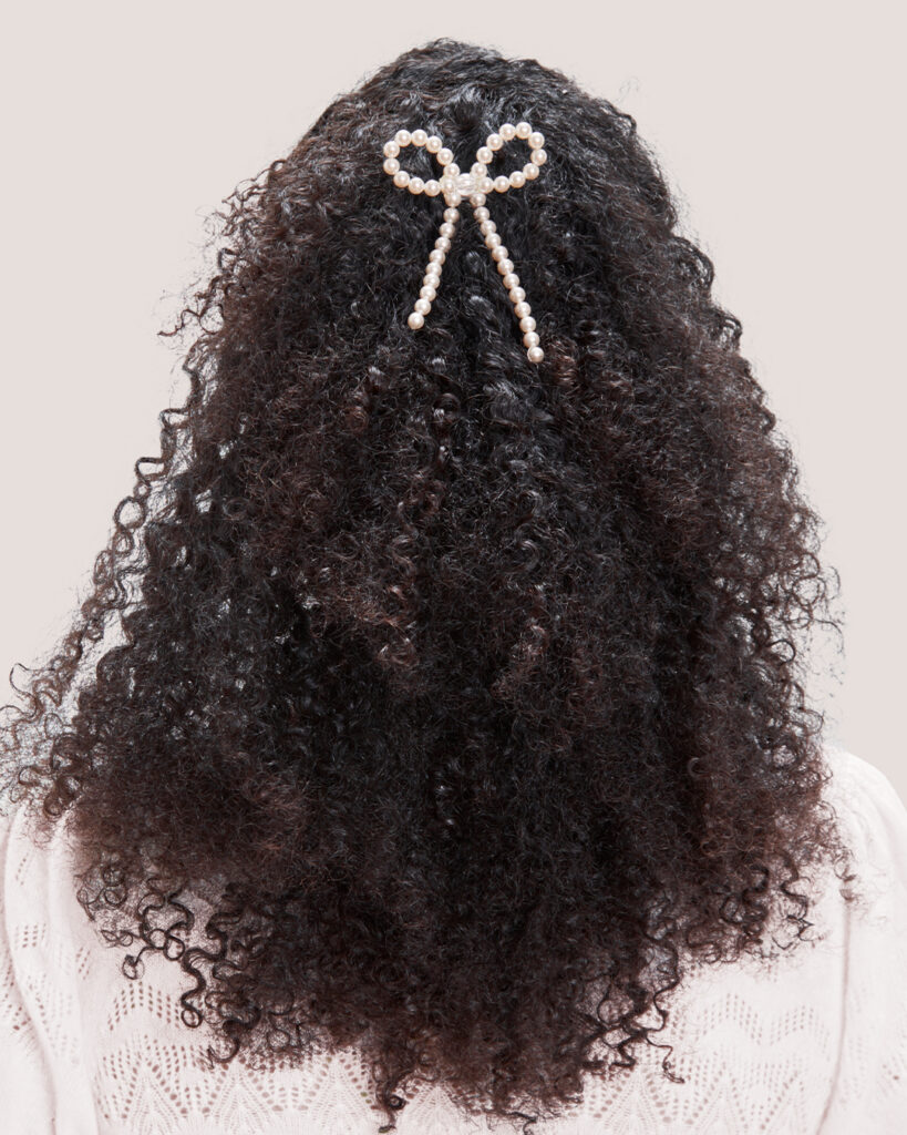 Curly hair styled half up with the Pearl Bow Bun Pin, viewed from the back against a plain background.