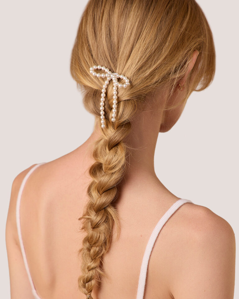 A woman with long blonde hair styled in a braid is pictured from behind, wearing the Pearl Bow Bun Pin.