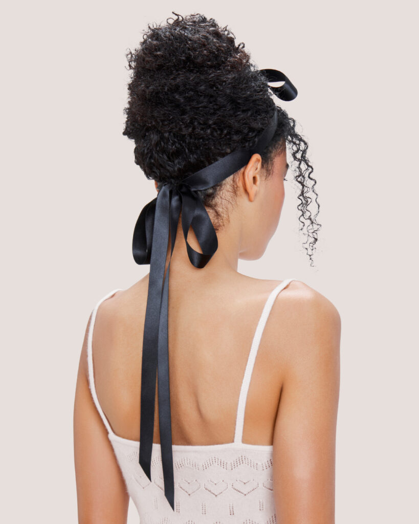 Woman with curly hair in a bun styled with the Black Satin Bow Hair Comb, paired with the HONOR Cashmere Heart Knit Camisole in Cream.