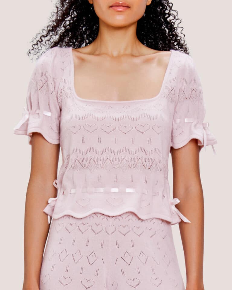 A woman wearing the Cashmere Heart Knit Top, a light pink, short-sleeved knit top featuring a square neckline and charming bow details on the sleeves.