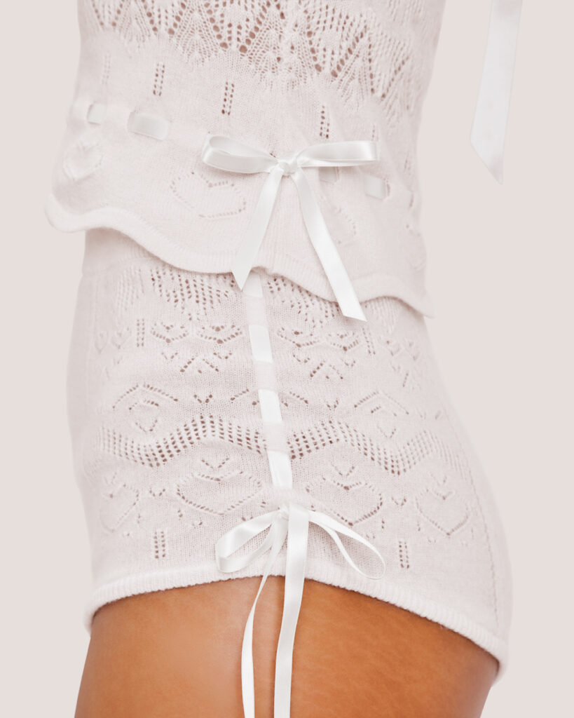 Close-up side view of the Cashmere Heart Knit Shorts in Cream, featuring knit fabric with satin ribbon bows, lace details, and a scalloped edge, photographed in front of a light background.