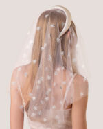 A woman with long blonde hair is shown from behind wearing the Embroidered Star Veil, a sheer white veil adorned with star patterns and paired with a White Padded Satin Headband.