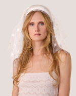 A woman with long blonde hair wears the Embroidered Star Veil—a white veil adorned with star patterns—the White Satin Padded Headband and the Cashmere Heart Knit Camisole in Cream.