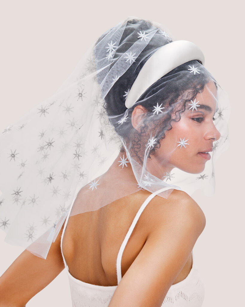 A woman in the Cream Cashmere Heart Knit Camisole wears a White Padded Satin Headband attached to the Embroidered Star Veil, looking to the side.