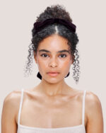 A woman with curly hair styled in a bun, facing forward, wears the Cashmere Knit Camisole in Cream and wears the Velvet Bow Hair Comb in Plum in her hair.