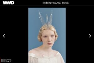 Woman in an off-shoulder white dress and clear swan headpiece poses against a blue background, as featured in a Womenswear Daily (WWD) "Bridal Spring 2027 Trends" article.