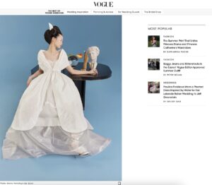Woman in a white gown leans on a black table in front of a blue background, featured in a Vogue weddings article.