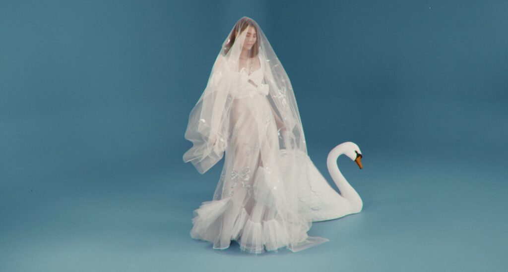 Woman in a sheer white dress and veil stands beside a white swan statue against a blue background.