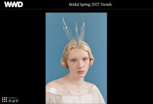 A model in a white dress wears a tall, clear, spiky headpiece against a blue background.
