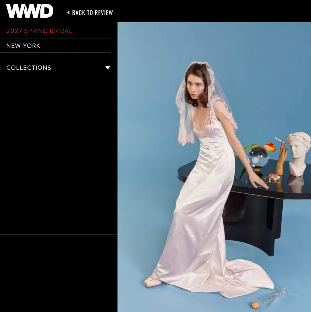 A woman in a satin bridal gown and veil poses by a black table with eclectic items, on a blue backdrop.