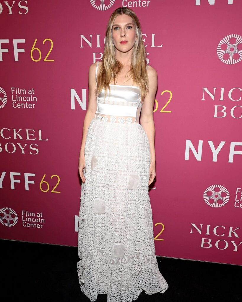 Woman in a white crop top and long lace skirt poses on a pink NYFF62 red carpet backdrop.