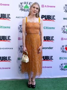 Woman in an orange lace dress holding a small bag, standing on a green carpet at an event backdrop.
