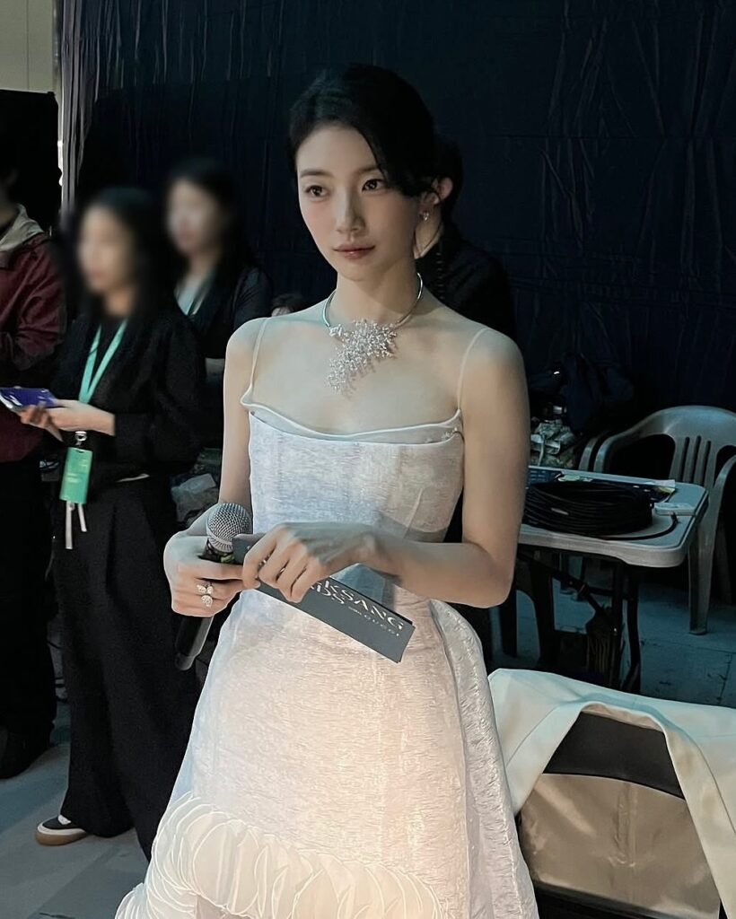 Woman in a white dress holding a microphone, standing backstage with people and chairs in the background.