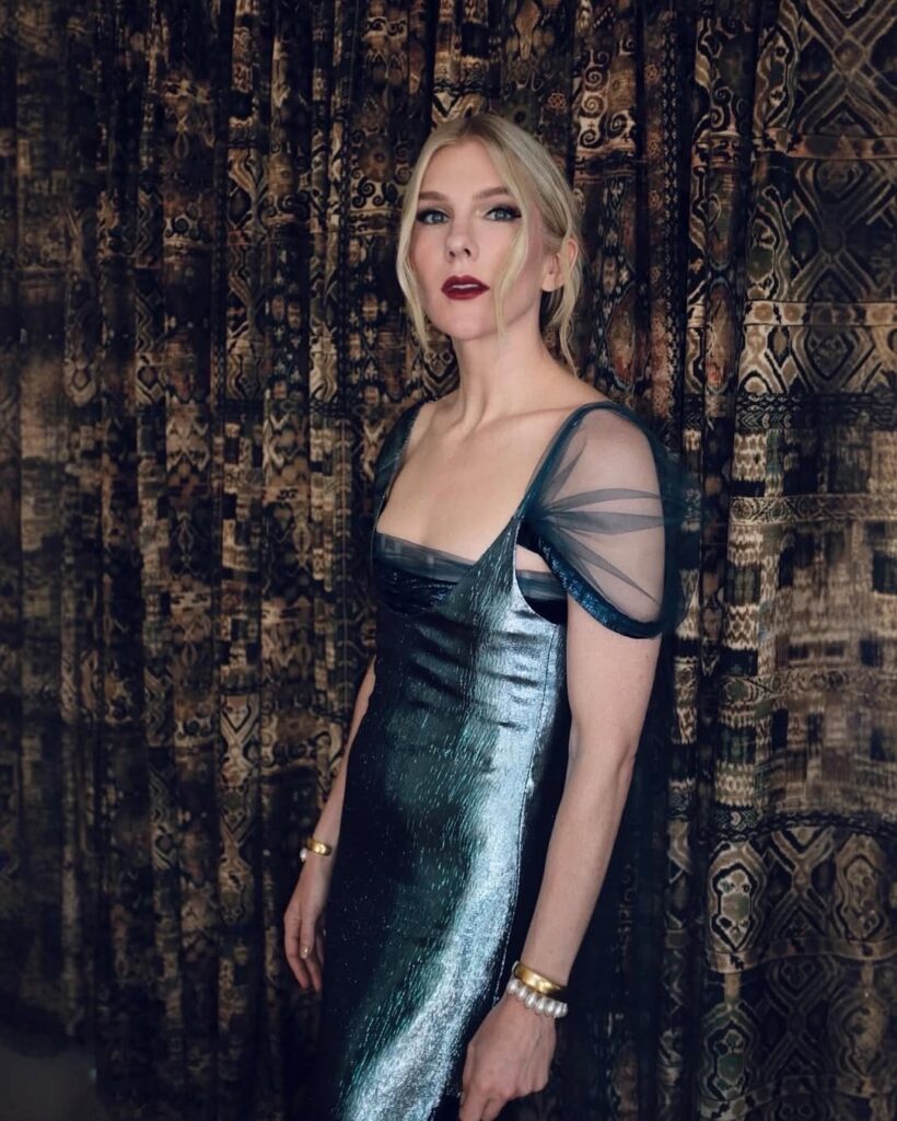 Woman in a metallic dress with sheer sleeves stands in front of patterned, dark curtains.