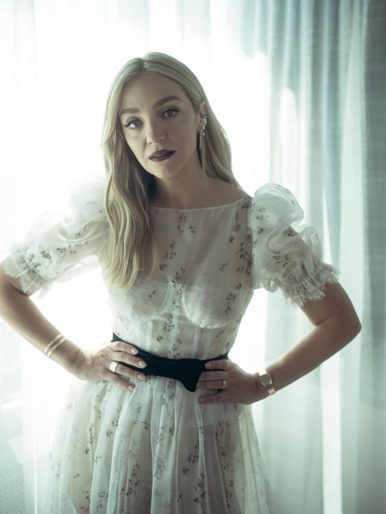 Woman in a sheer white floral dress standing in front of a curtain, hands on hips, looking at camera.