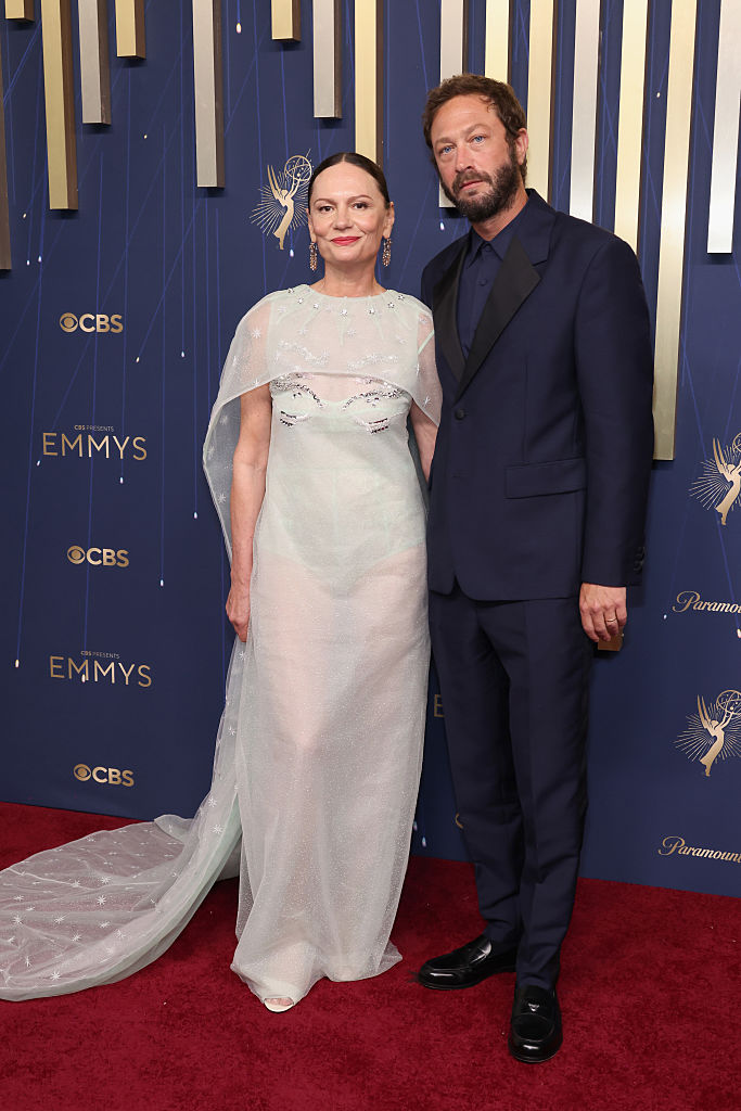Two people posing on a red carpet at the Emmys, one in a sheer gown and the other in a dark suit.