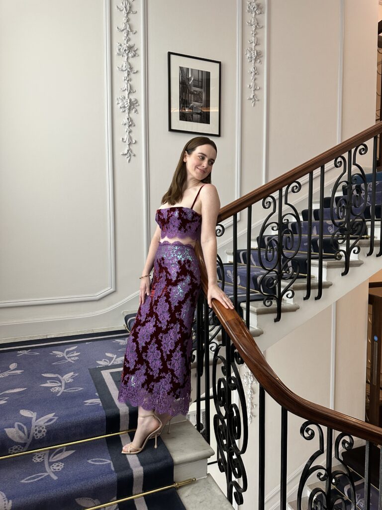 Woman in a purple lace dress stands on a carpeted staircase with ornate railings, smiling at the camera.