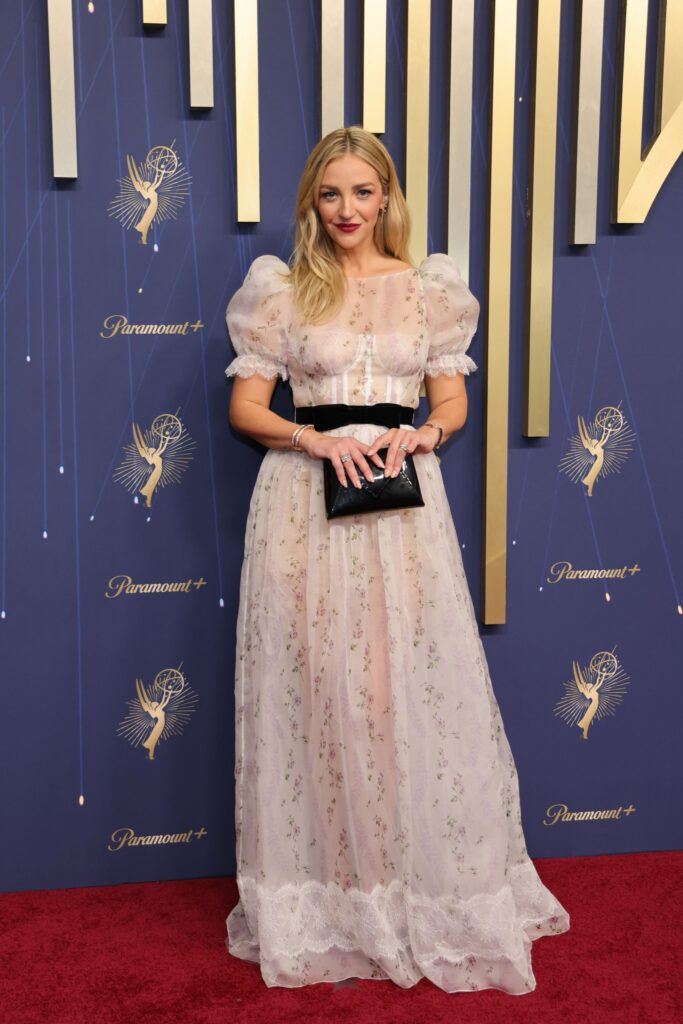 A woman in a sheer, puff-sleeve floral gown holds a black clutch on a red carpet with Emmy logos behind her.