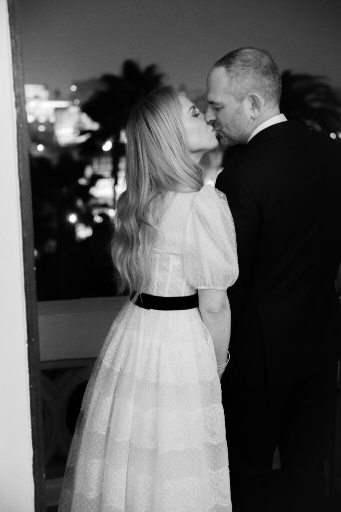 A woman in a white dress kisses a man in a suit on a balcony at night.